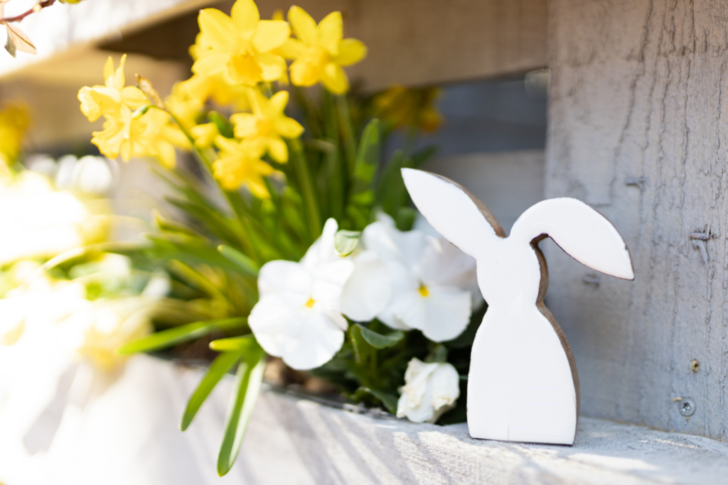 Eine weiße Hasenfigur steht auf einem Regel vor einem Strauß Narzissen.