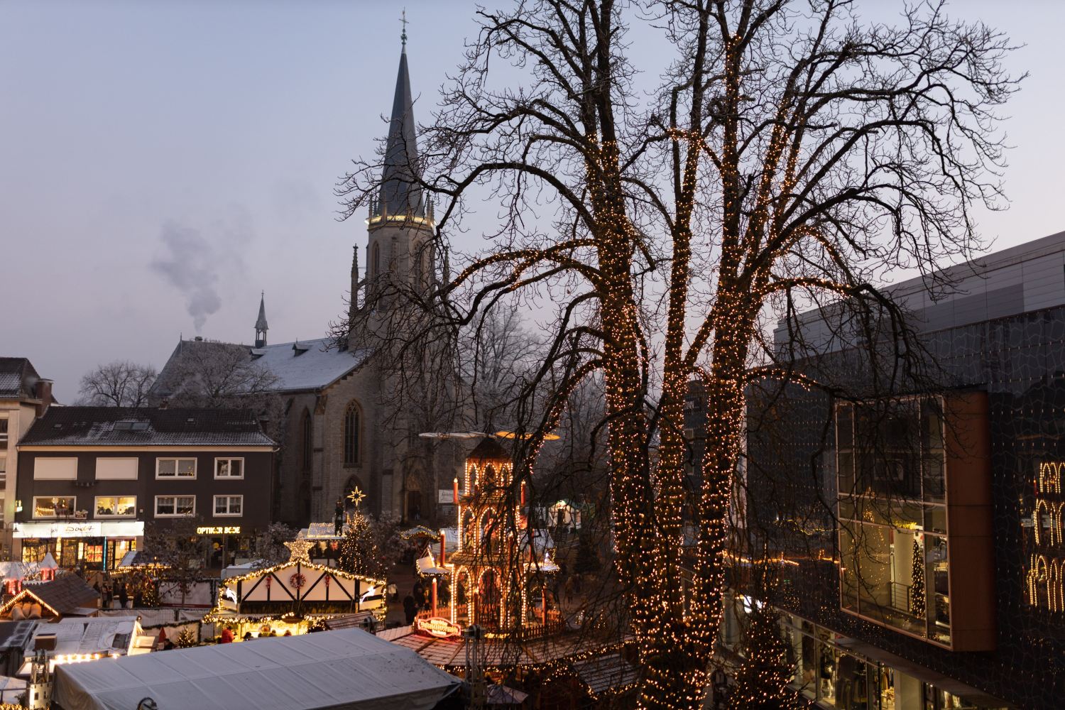 Der Weihnachtsmarkt auf dem Berliner Platz in Gütersloh bei Abenddämmerung. Im Hintergrund die Martin-Luther-Kirche. Vorne eine großer Baum mit Lichterkette sowie weihnachtlich geschmückte Hütten.