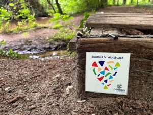 Vorne eine Plakette mit bunten Schnipseln und der Überschrift Stadtteil Schnipsel-Jadg sowie dem Jubiläumslogo, im Hintergrund ein Bachlauf und Bäume
