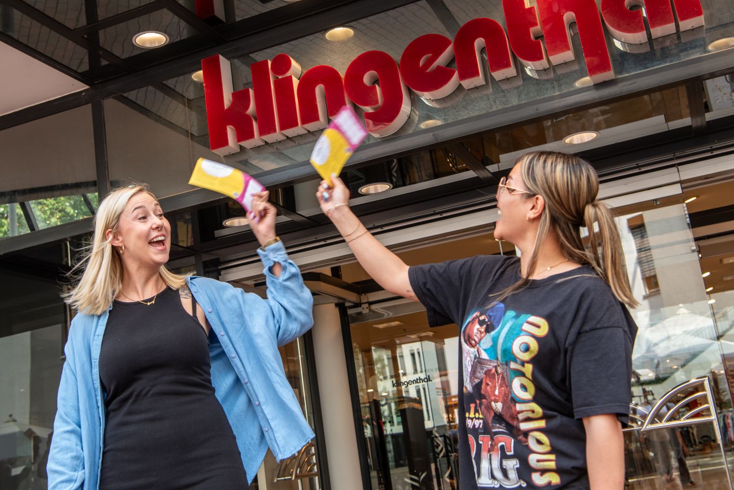 Zwei junge Frauen klatschen sich mit einer Karte in den Händen ab. Sie lächeln und stehen vor einem Geschäft.