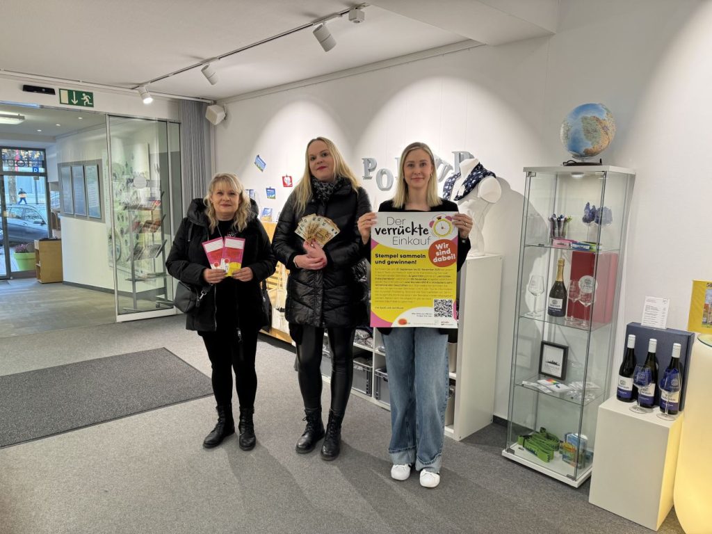 Drei Frauen schauen in die Kamera, sie halten Flyer und ein Plakat vom "Verrückten Einkauf". Die Frau in der Mitte hält Geldscheine im Wert von 500 Euro in der Hand.