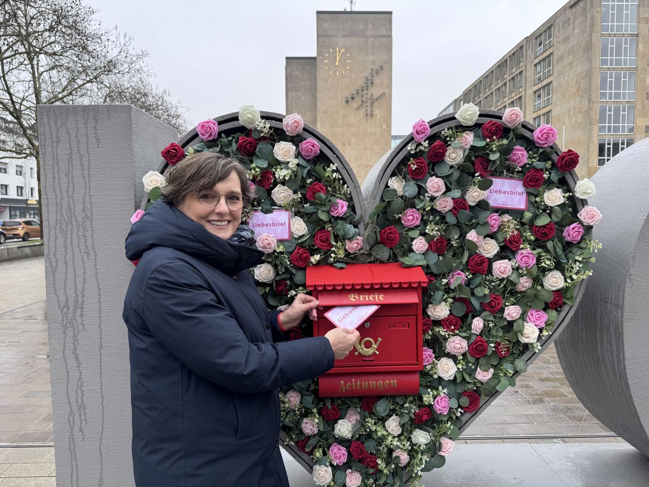 Liebesbriefe an Gütersloh 1 © Stadt Gütersloh Eine Frau, Wiebke Heine, wirft eine Postkarte in einen roten Briefkasten, der inmitten eines mit Blumen geschmückten Herzens eingebaut ist. Sie schaut in die Kamera und lächelt.