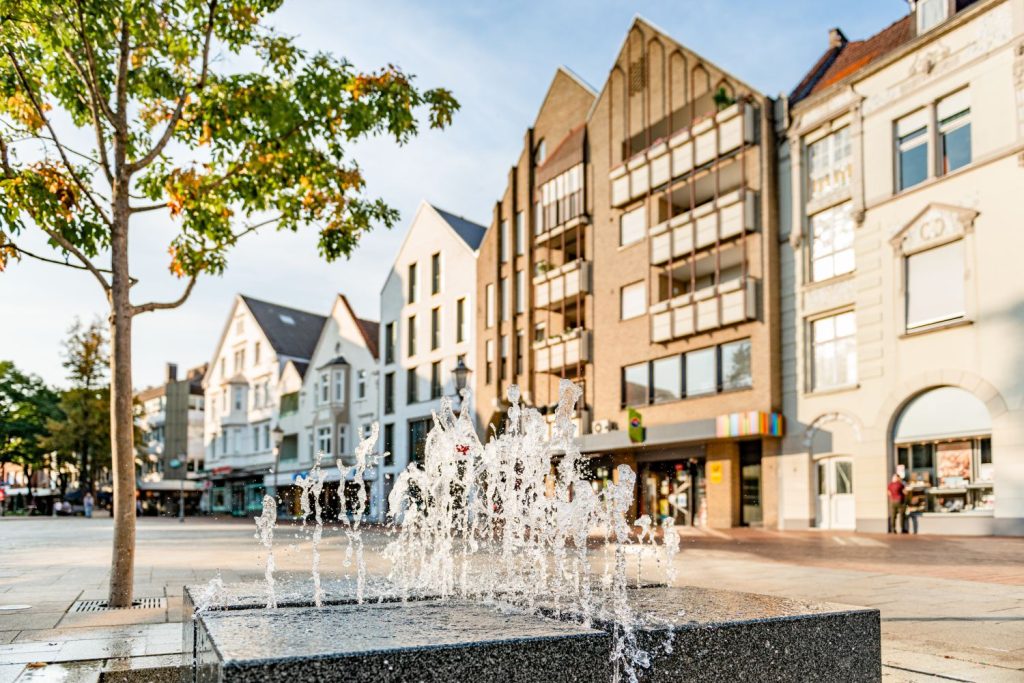 Ein Wasserspiel vor einer Innenstadt-Häuserzeile bei gutem Wetter.