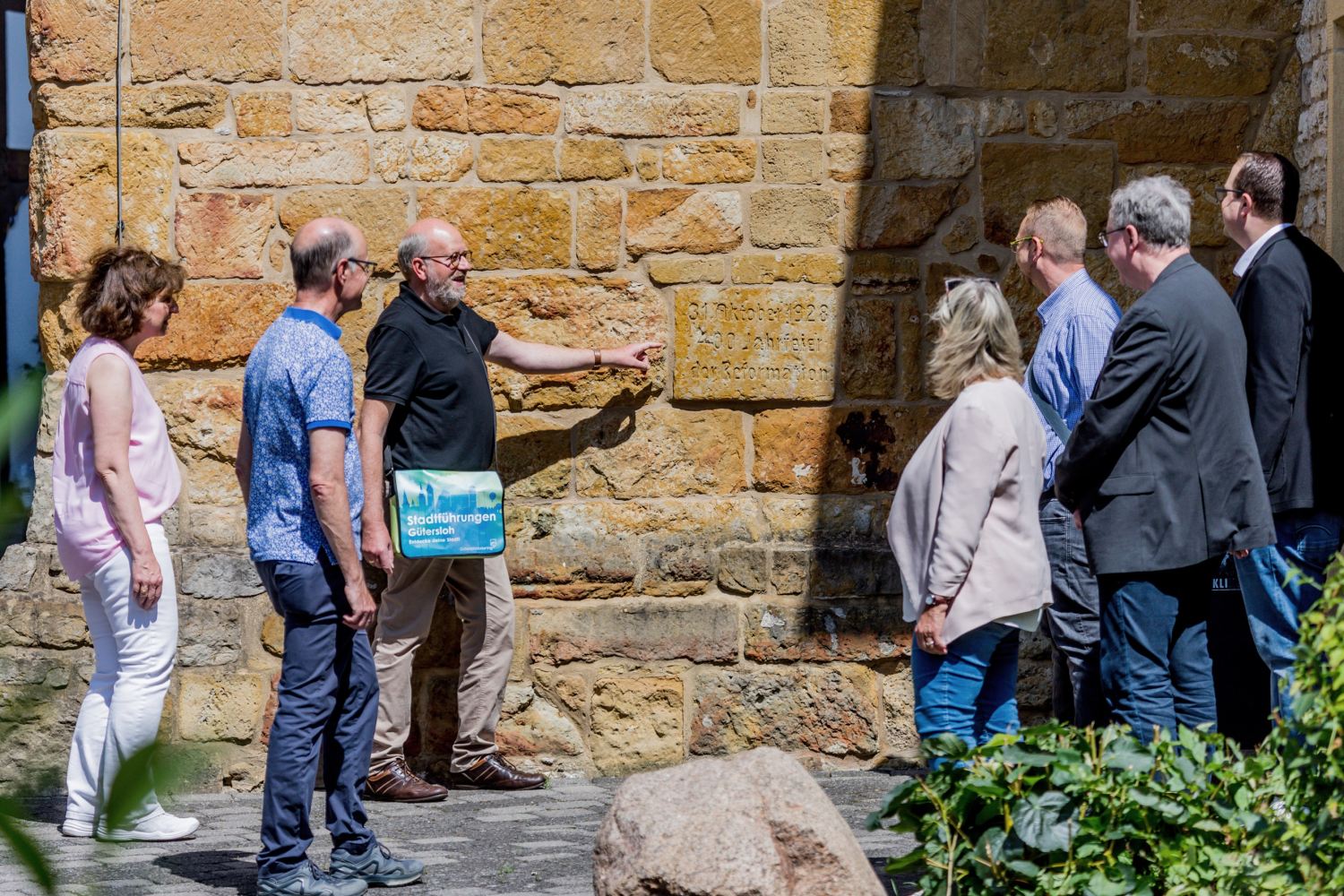 Sieben Personen stehen um eine an einer Wand angebrachten Plakette. Ein Mann zeigt darauf und spricht, er trägt eine Tasche mit der Aufschrift "Stadtführungen Gütersloh"