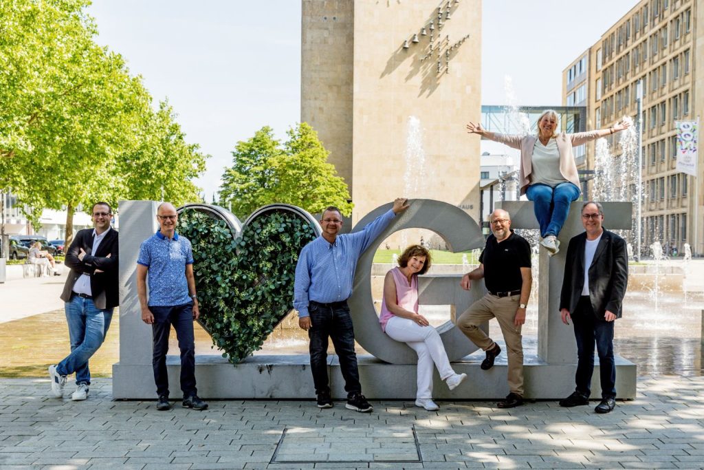 Sieben erwachsene Personen stehen und sitzen um, am und auf einem mannshohen Schriftzug aus Beton. Sie schauen lächelnd in die Kamera. Der Schriftzug lautet "I bepflanztes Herz GT". Im Hintergrund ist das Rathaus Gütersloh zu sehen sowie einige Bäume, ein Wasserspiel.