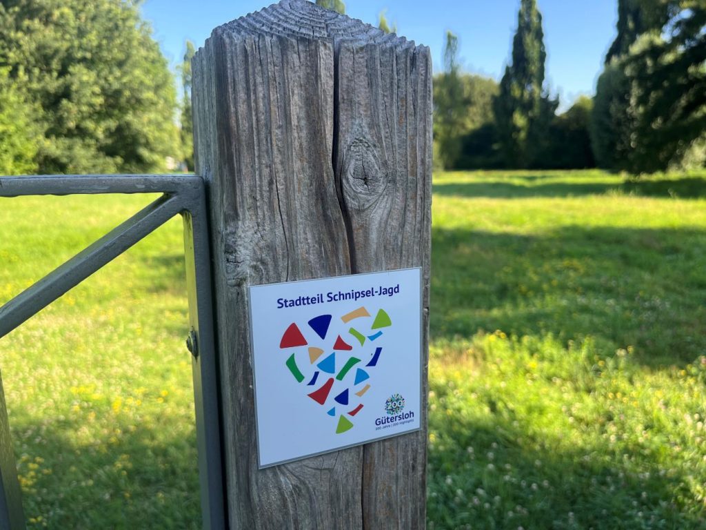 Ein Schild an einem Holzzaun mit der Aufschrift 'Stadtteil Schnipsel-Jagd' und bunten geometrischen Formen.