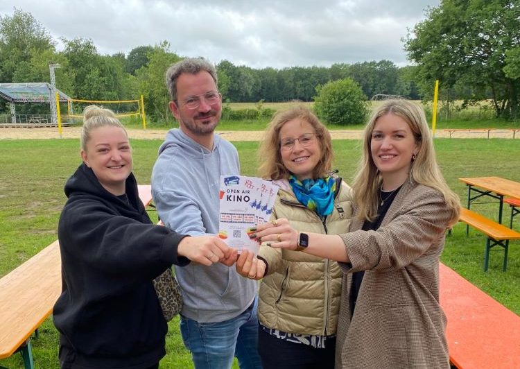 Vier Personen stehen im Freien auf einer Wiese und halten gemeinsam einen Flyer mit der Aufschrift „OPEN AIR KINO“ in die Kamera.