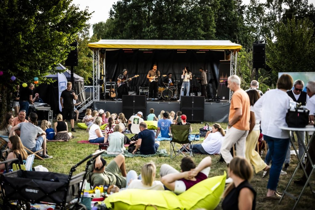 Menschen sitzen auf einer Wiese vor einer Bühne bei einem Live-Konzert im Freien.