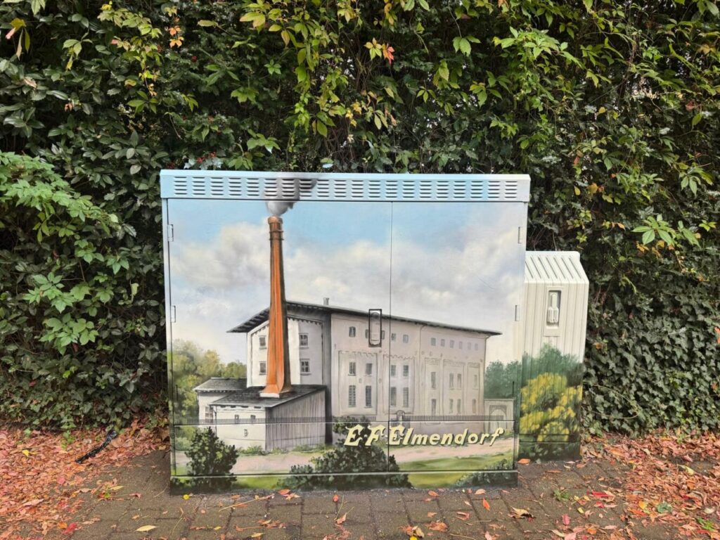 Ein bemalter Verteilerkasten zeigt ein Gebäude mit Schornstein und die Aufschrift „E.-F.-Elmendorf“ vor dichtem Grün.